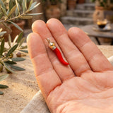 Hand holding a red chili pepper-shaped earring with a gold and silver top, outdoor setting.